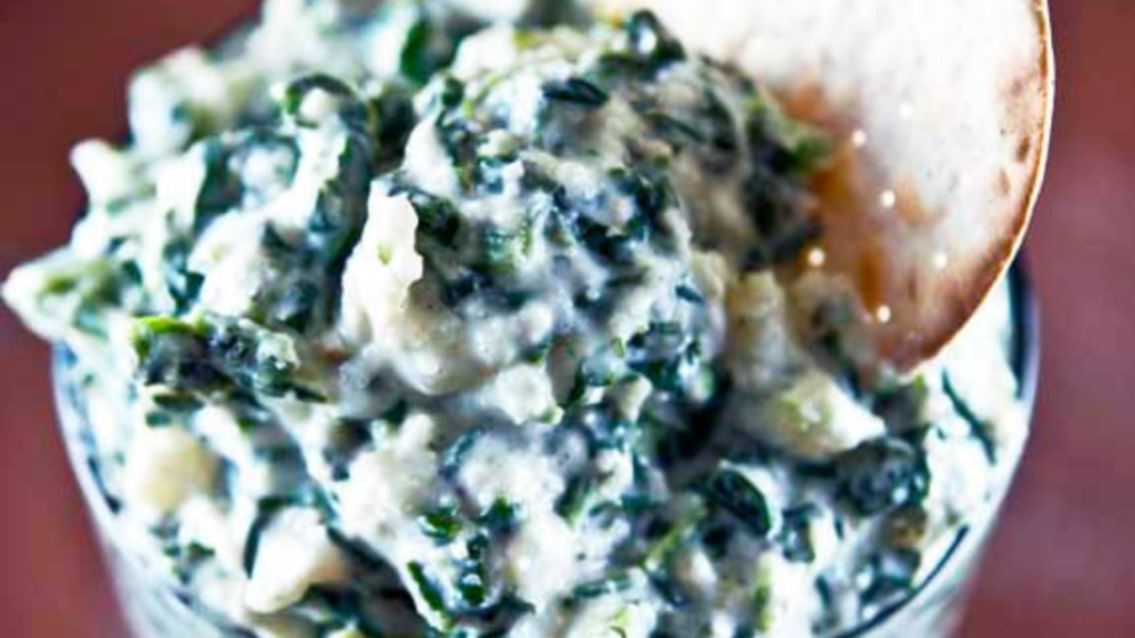 A closeup of a bowl holding spinach dip. A cracker is nestled into the dip.