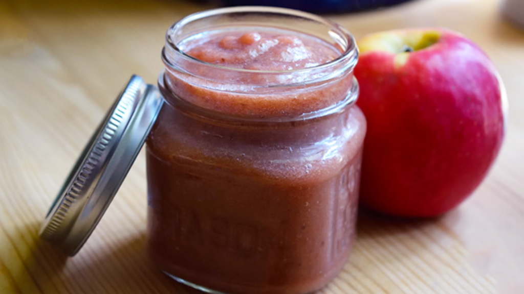 A small mason jar full of strawberry apple sauce. An apple sits next to the jar.