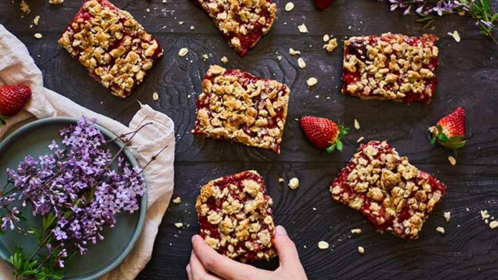 A hand reaches for a strawberry crumble bar from a wood surface where several bars are spread out.