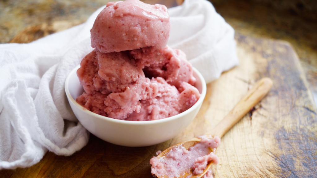 A small white bowl sits filled with strawberry n'ice cream. A used wooden spoon sits in front of the bowl.