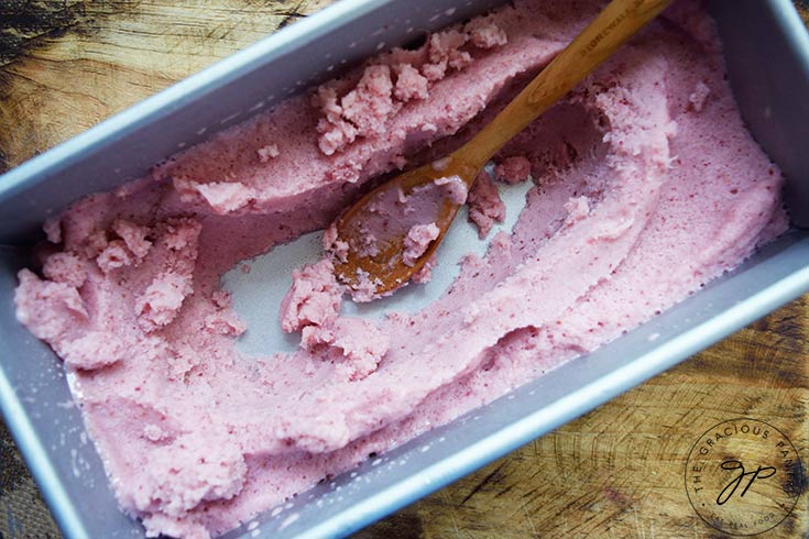 A loaf pan of Strawberry N'ice Cream with some of the Strawberry N'ice Cream dug out of the middle. A spoon rests in the hole.