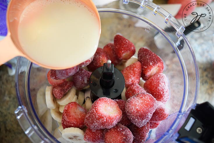 Pouring milk into frozen strawberries and bananas in a food processor.