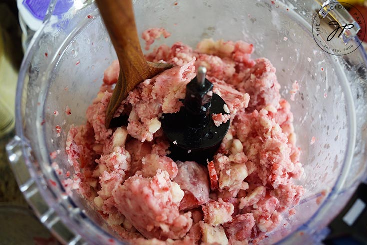 Partially blended Strawberry N'ice Cream in a food processor. A wood spoon tamps down some of the blended fruit.