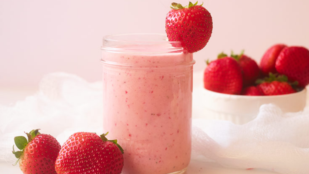 A glass filled with strawberry smoothie. Fresh strawberries lay around the glass.