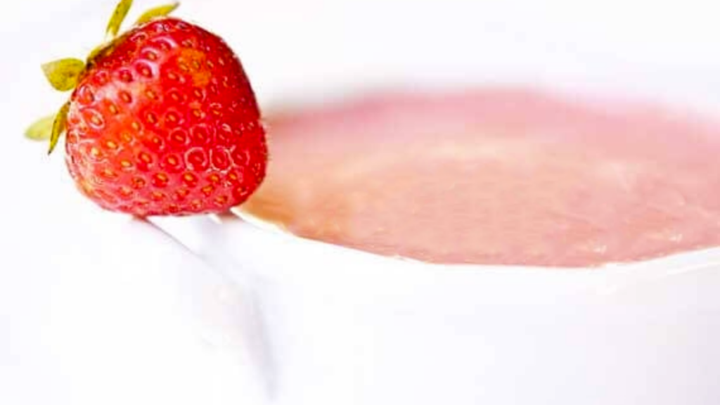 A white bowl holds a serving of strawberry soup. A fresh strawberry rests on the handle.