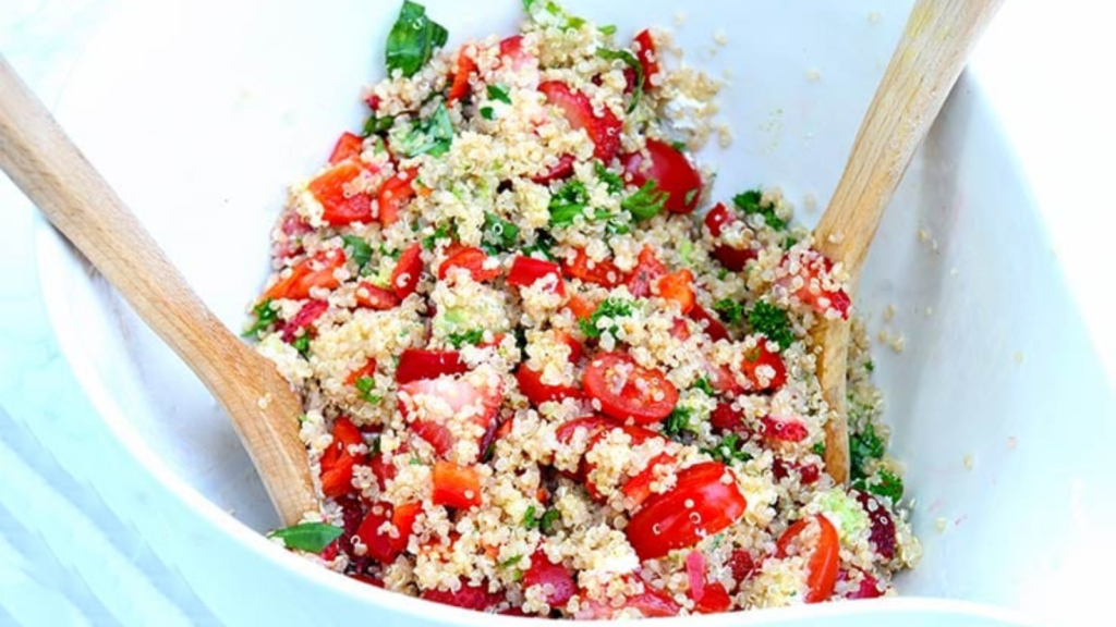 A white mixing bowl holds summer quinoa salad and two wooden spoons.