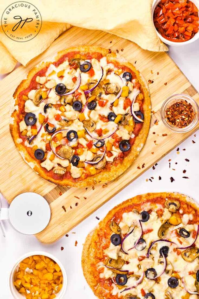 An overhead view of a Supreme Pizza on a cutting board and another next to it on a white surface.