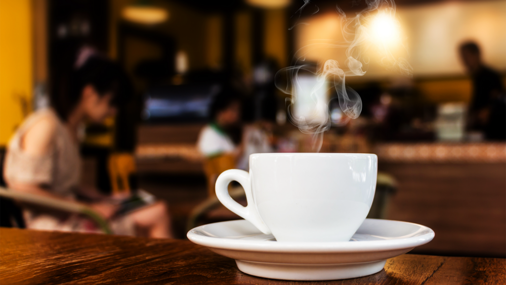 A white cup and saucer with steam coming up sits on a table in a coffee shop.