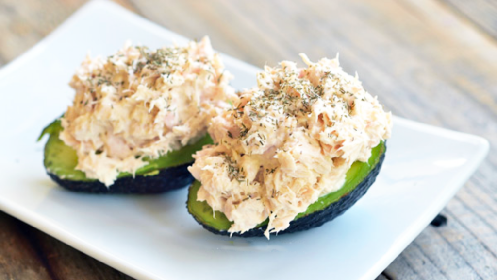 Two avocado halves lay on a white plate and are both stuffed with tuna salad.