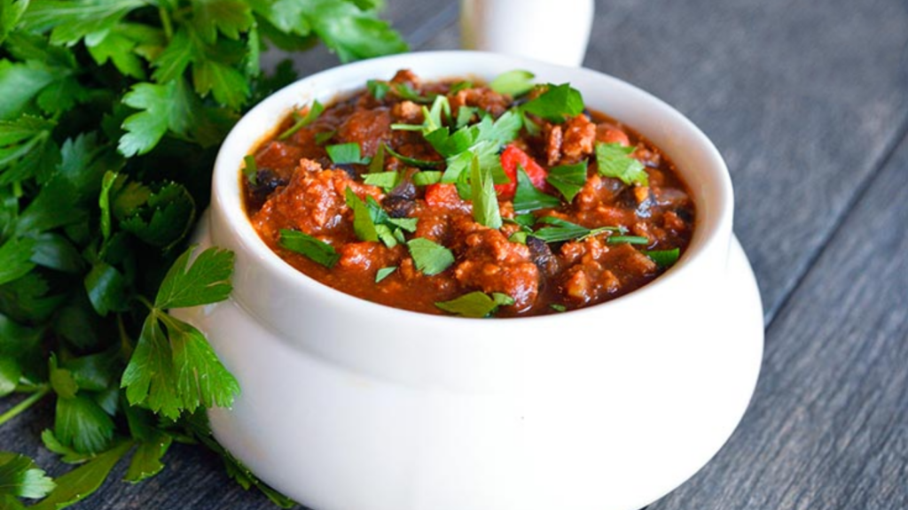 A white crock filled with turkey chili and garnished with fresh parsley.