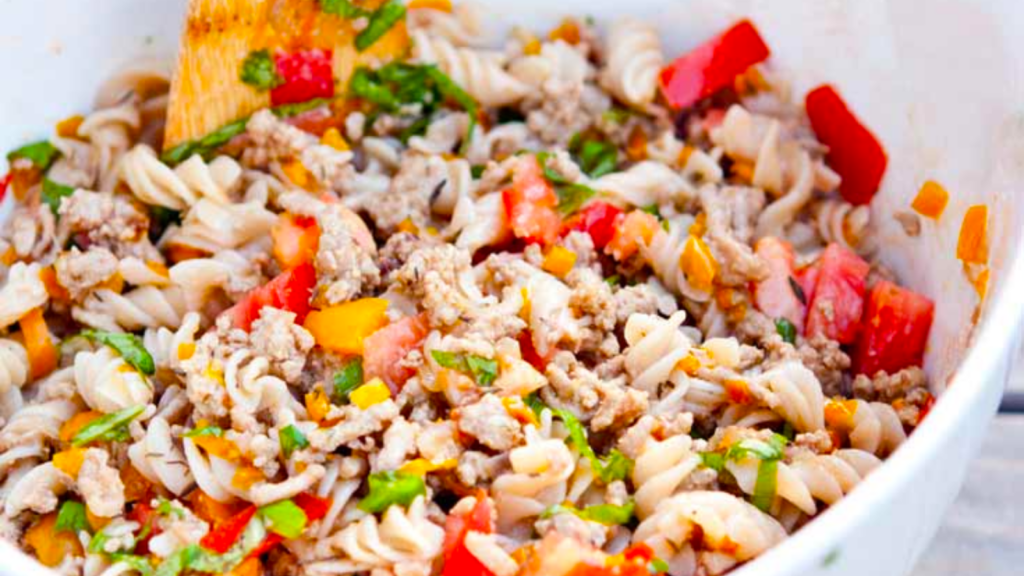 A white mixing bowl sits filled with turkey vegetable pasta salad.