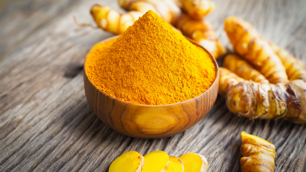 A bowl of dry, ground turmeric sits surrounded by fresh turmeric.