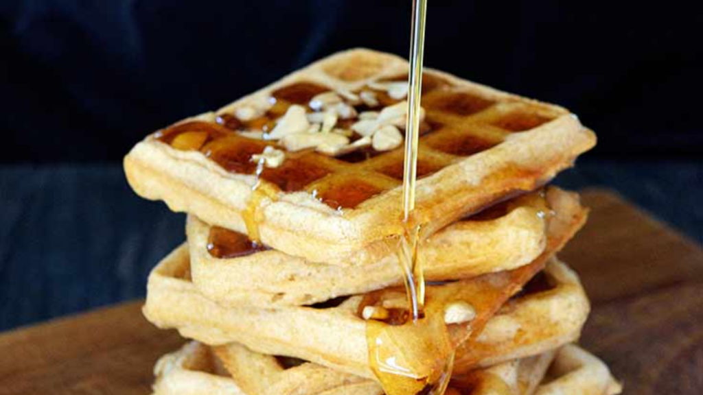 A stac of vegan waffles with syrup pouring down over them and nuts sprinkled on top for garnish.