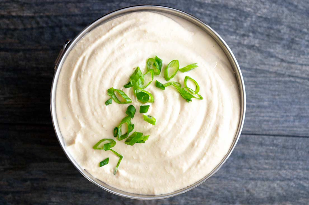 An overhead view of a metal serving bowl filled with white bean hummus and garnished with sliced green onions.