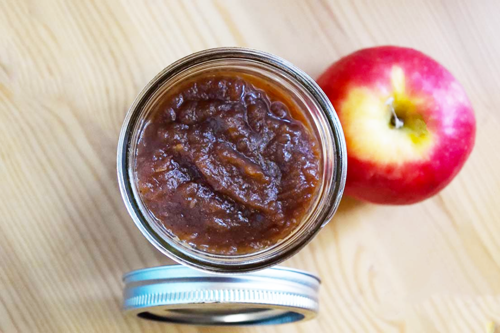 An overhead view of an open jar filled with amish apple butter. The jar lid and an apple lay to the sides of the jar.