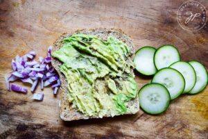 A slice of avocado toast laying in between a pile of chopped, red onions and slices of cucumber.