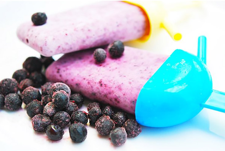 Two frozen blueberry popsicles laying on a white surface with a few frozen blueberries.