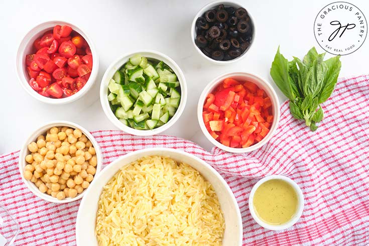 Chickpea Orzo Salad recipe ingredients gathered in individual white bowls on a white surface.