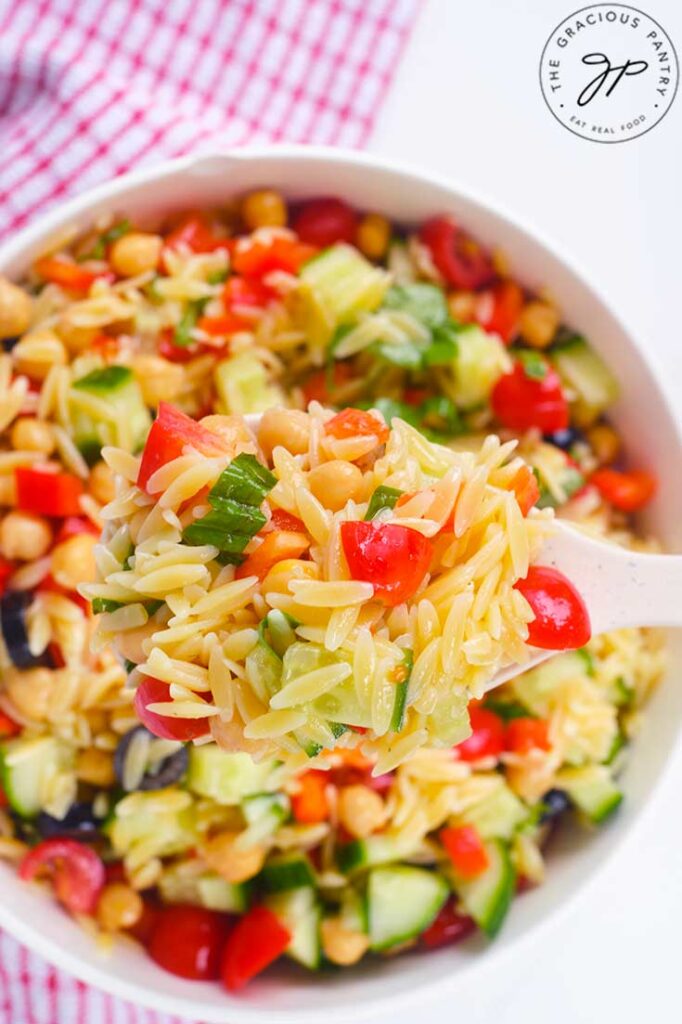 A white spoon lifts some Chickpea Orzo Salad out of a white bowl.