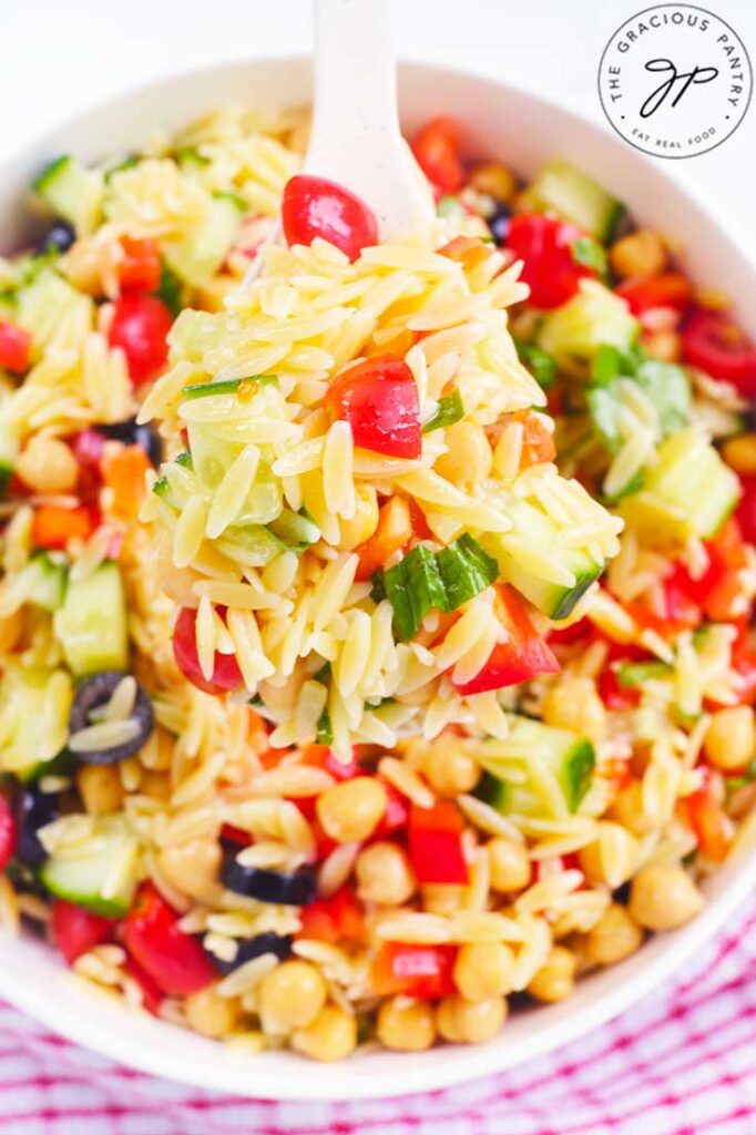 A white spoon lifts some Chickpea Orzo Salad out of a white bowl filled with it.