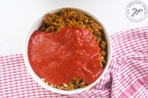 Marinara added to pasta and ground turkey in a white mixing bowl.