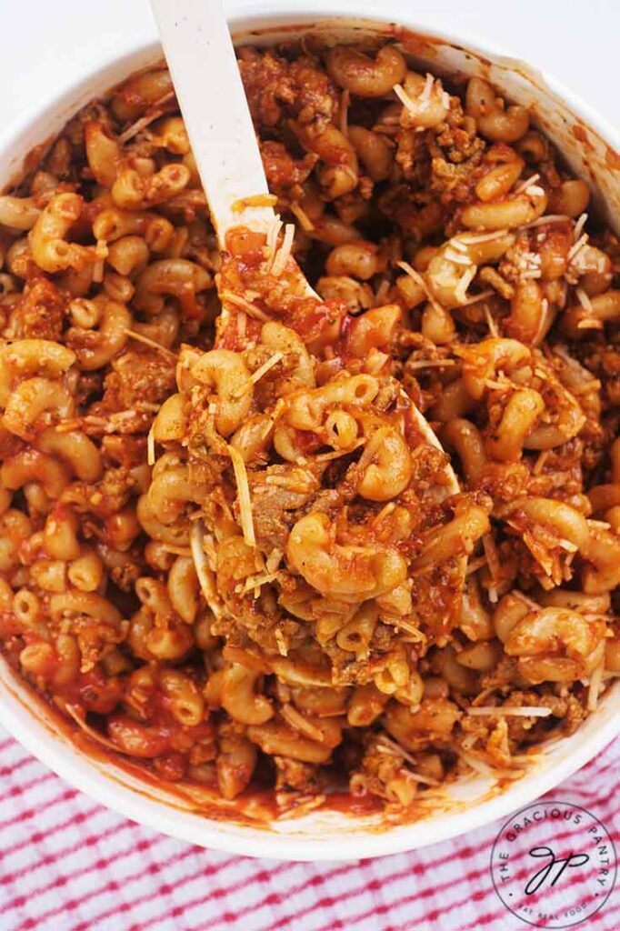 A spoon lifts a spoonful of chili mac up out of a white bowl filled with it.