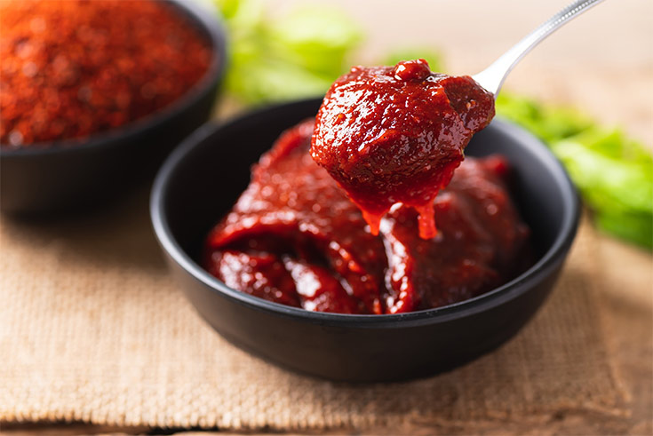 A small black bowl filled with Gochujang. A spoons lifts up a spoonful of it.