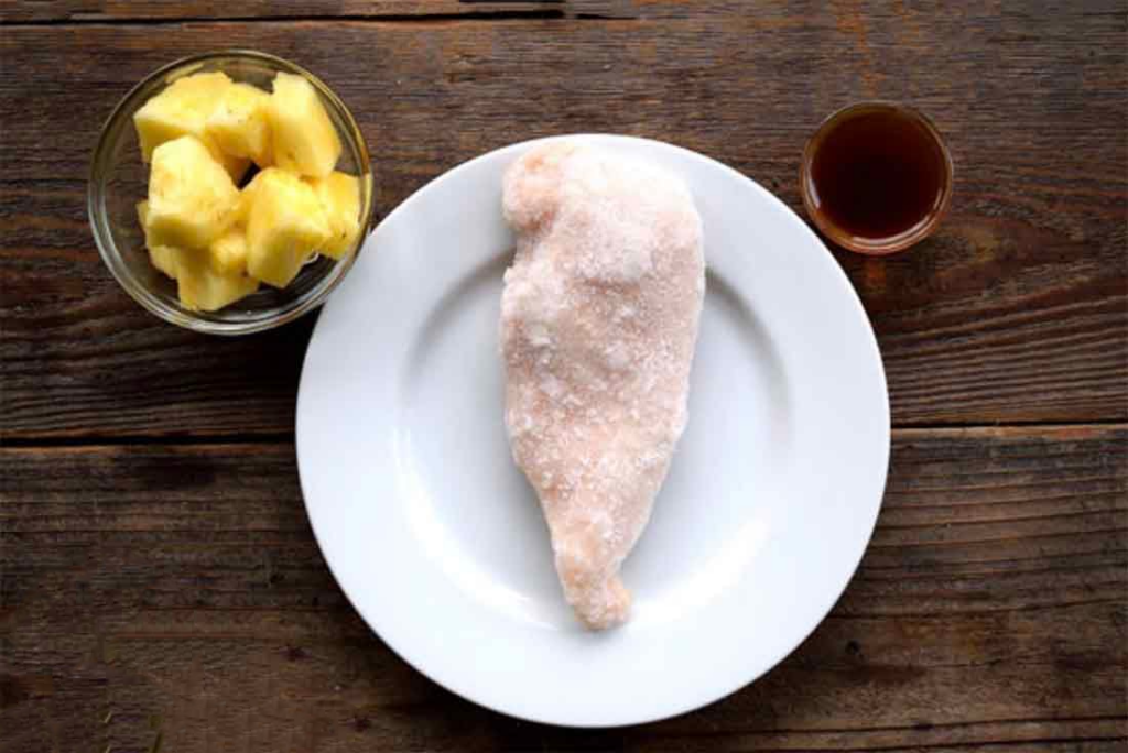 A white plate with a frozen chicken breast sits between 2 glass bowls. One with sauce and the other with pineapple chunks.