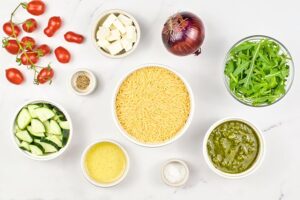 Ingredients for Orzo Salad With Pesto in individual bowls on a white surface.