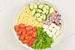 Chopped cucumber added to orzo in a white mixing bowl.