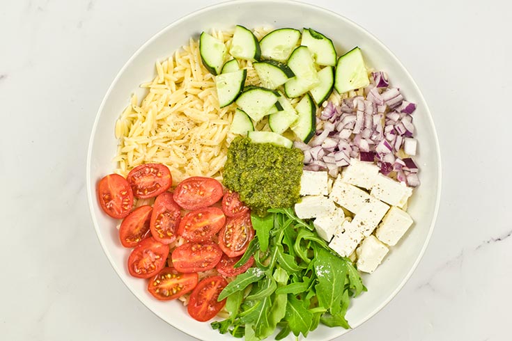 Ground pepper added to Orzo Salad With Pesto Recipe ingredients in a white mixing bowl.