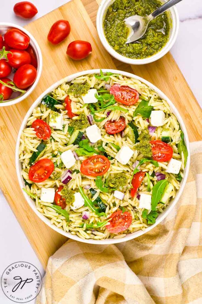 An overhead view of a white bowl sitting on a cutting board, filled with Orzo Salad With Pesto.