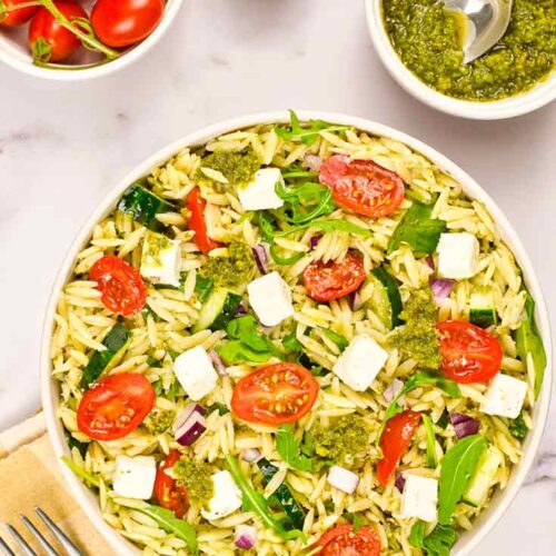 A white bowl filled with Orzo Salad With Pesto. Small bowls of tomatoes and pest sit to the side.