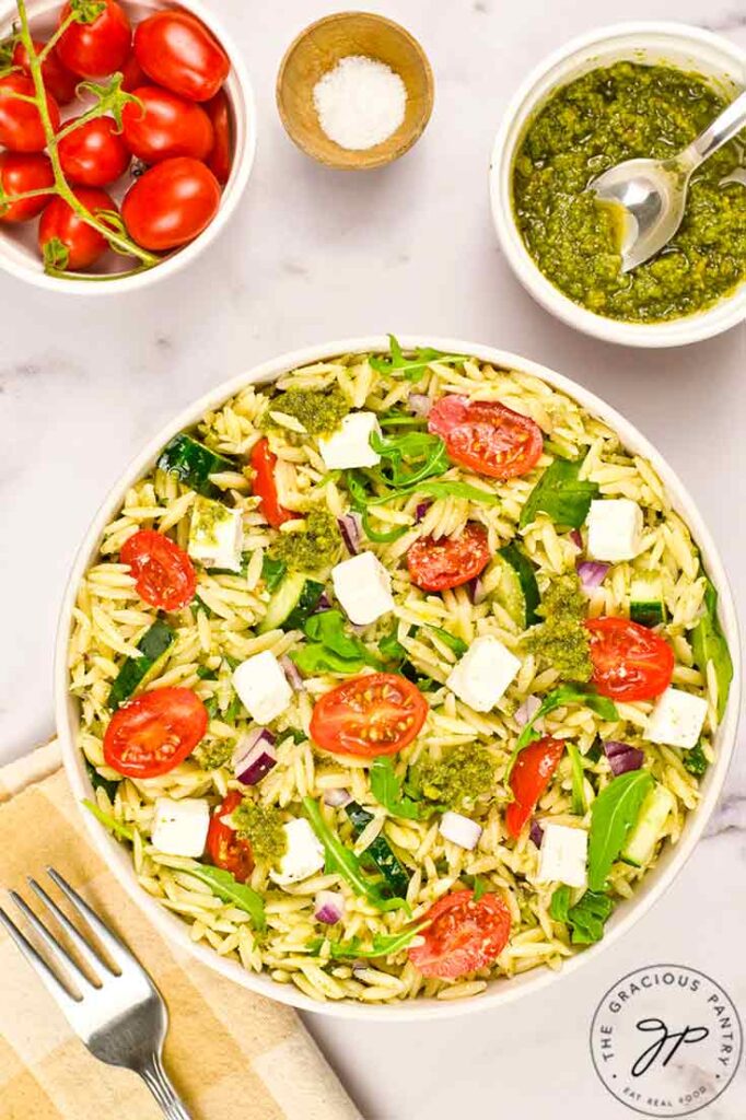 A white bowl filled with Orzo Salad With Pesto. Small bowls of tomatoes and pest sit to the side.