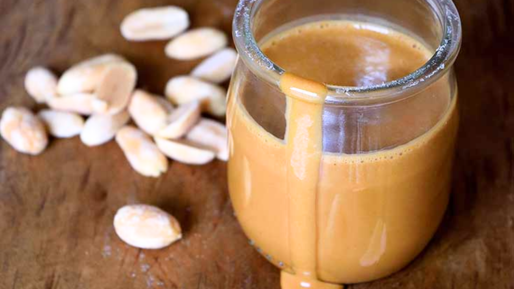 A small, glass jar filled with peanut sauce sits on a wood surface.