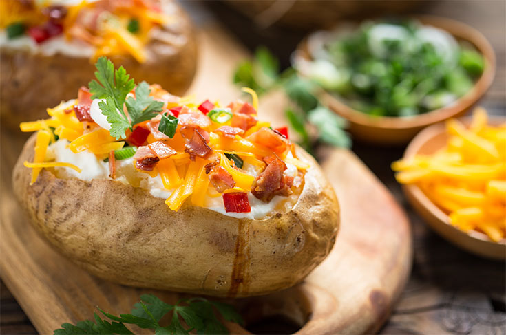 A baked potato topped with grated cheddar cheese and bacon bits sits on a cutting board.