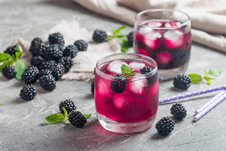 Two glasses with blackberry lemonade surrounded by fresh blackberries and mint leaves.