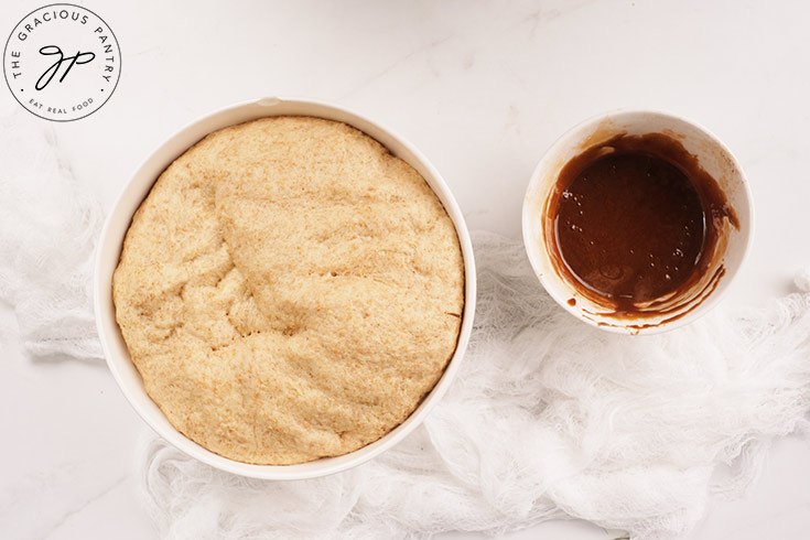 Risen dough in one bowl and honey glaze in a second bowl on a white surface.