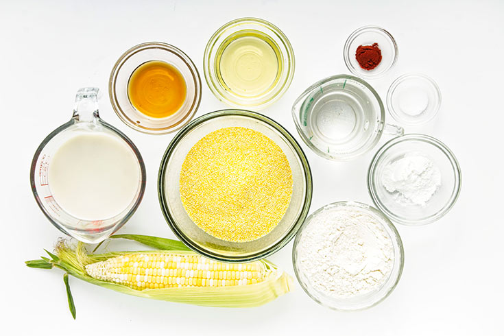 Cornbread muffin ingredients in individual bowls on a white surface.