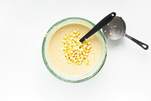 Corn kernels added to cornbread muffin batter in a glass mixing bowl.