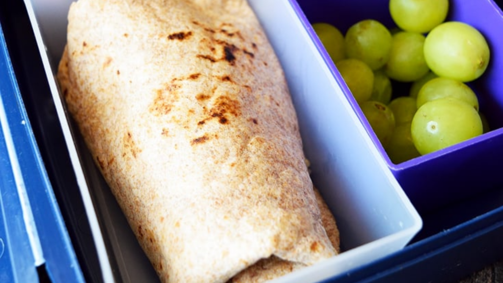 A burrito in a bento container next to a second container of green grapes.