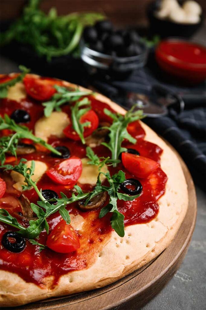 A pita pizza on a round cutting board, topped with tomato sauce, tomatoes and arugula.