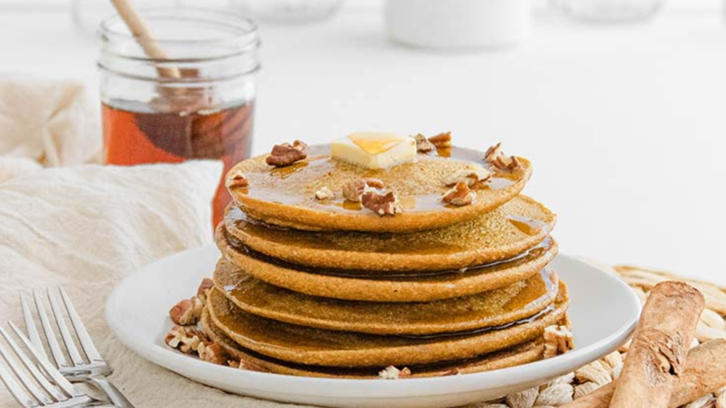 A stack of Pumpkin Oatmeal Pancakes on a white plate, topped with butter, syrup and pecans.