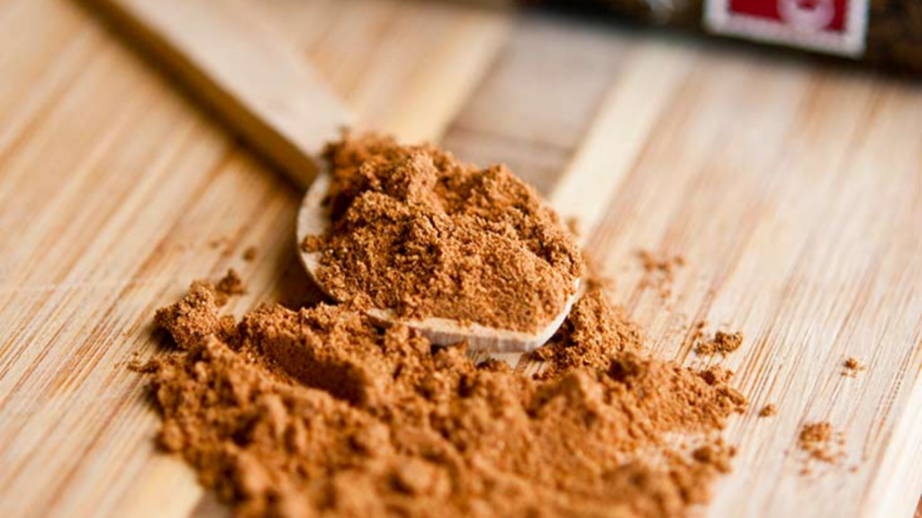 A small wooden spoon filled with pumpkin pie spice lays in a pile of the same spice on a wooden surface.