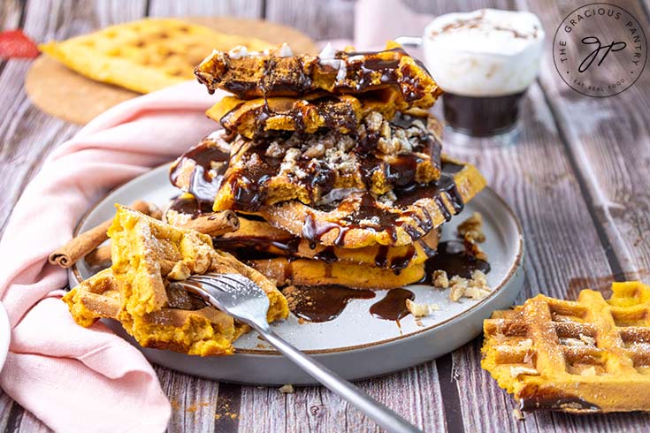 A large stack of pumpkin waffles served on a plate with chocolate syrup and whipped cream over the top.