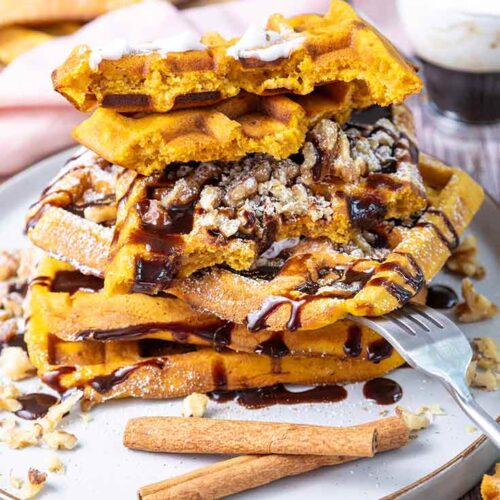 A large stack of Pumpkin Waffles on a plate topped with whipped cream and drizzled with chocolate syrup.