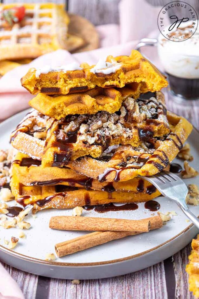 A large stack of Pumpkin Waffles on a plate topped with whipped cream and drizzled with chocolate syrup.
