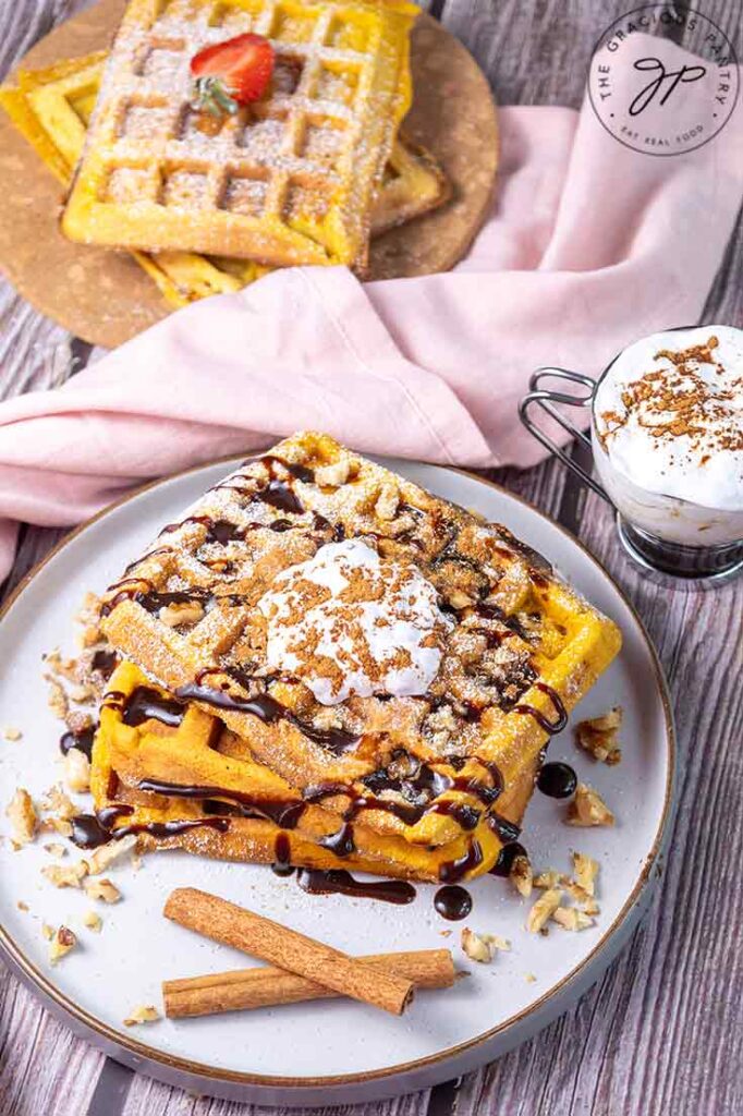 A slightly overhead view of a stack of uncut pumpkin waffles on a plate. They are garnished with whipped cream, cinnamon, and chocolate syrup.