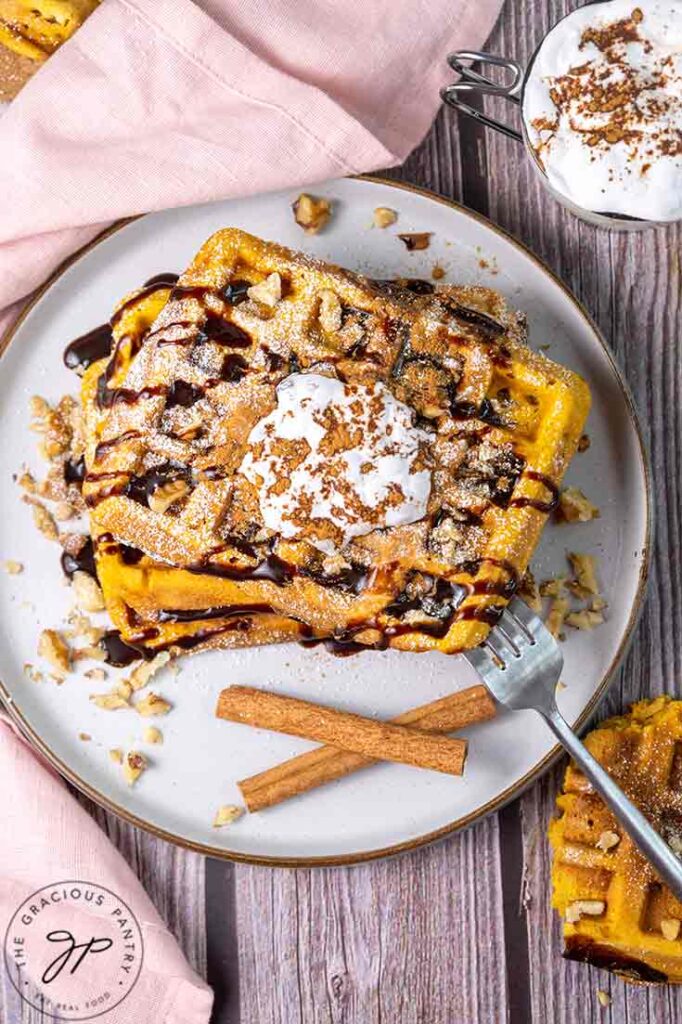 An overhead view of a stack of Pumpkin Waffles on a plate topped with whipped cream, ground cinnamon and chocolate syrup.