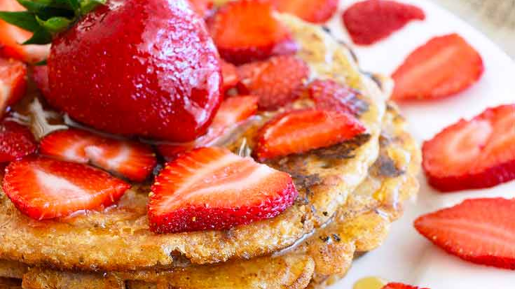 A short stack of spelt pancakes covered in strawberry slices and maple syrup.